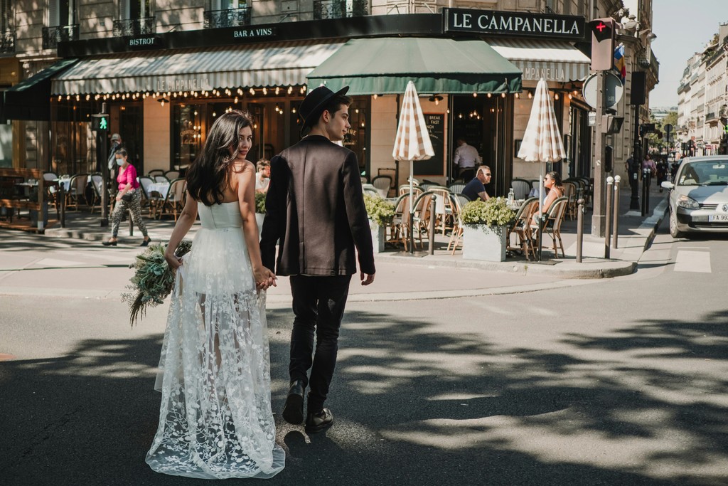 Bride glancing back while walking hand in hand with the groom past a charming Parisian sidewalk cafe on a sunny day