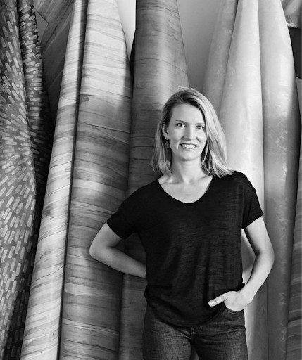 White woman with blonde hair posing in front of large rolls of fabric.