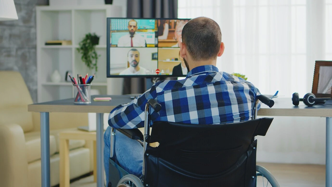 A imagem mostra um homem sentado em uma cadeira de rodas, participando de uma videoconferência. Ele está de costas para a câmera, usando uma camisa xadrez azul e branca, e está de frente para uma mesa com um monitor que exibe a tela dividida em quatro, com quatro pessoas participando da chamada. O ambiente parece ser um escritório em casa, com estantes ao fundo e itens como fones de ouvido e materiais de escritório sobre a mesa. A imagem transmite um ambiente de trabalho remoto ou de recrutamento online, destacando a acessibilidade e inclusão de pessoas com deficiência no mercado de trabalho.