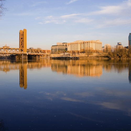 在日落时分，在晴朗的天空下，一条平静的河流倒映着建筑物和一座桥。