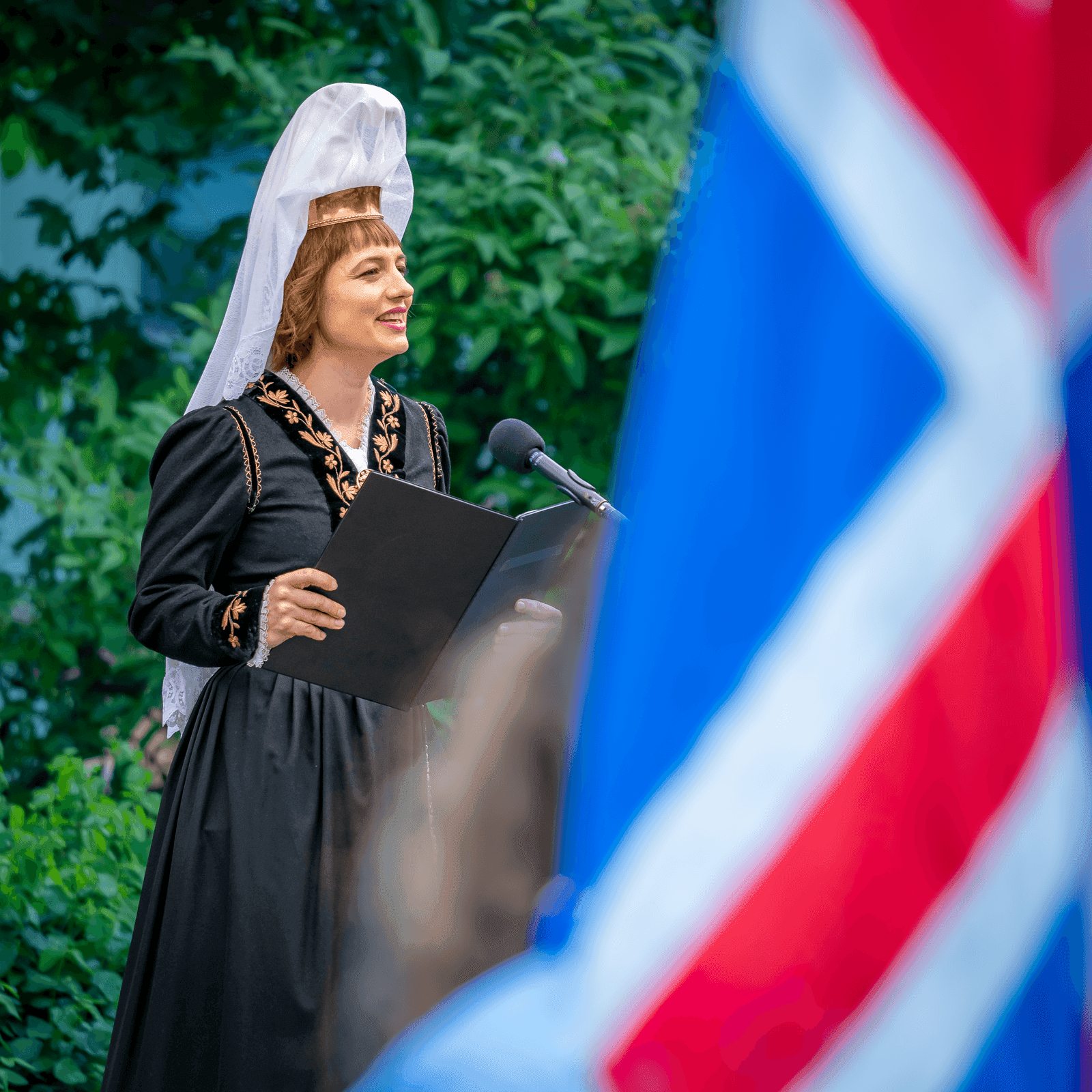 A woman representing the Fjallkona, the Lady of the Mountain, addresses the crowd at a microphone during Iceland's National Day celebrations on June 17. She wears the traditional black Icelandic costume with gold embroidery and a white lace headdress, with a blurred Icelandic flag in the foreground.