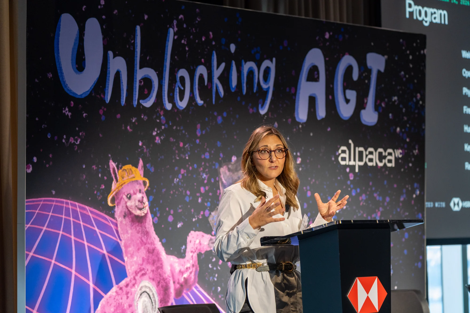 Aubrie Pagano speaking at the HSBC lectern, with the Unblocking AGI and Alpaca backdrop behind her.