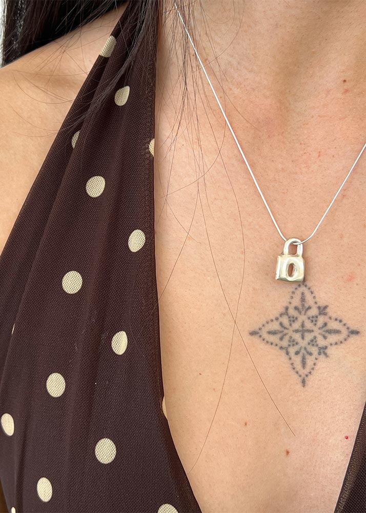 Close-up of a person wearing a silver necklace with a lock pendant, featuring a tattoo on the skin.