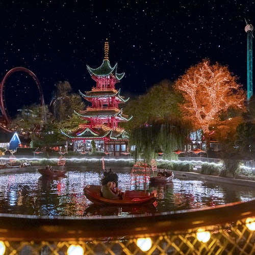 En oplyst pagode og træ spejler sig i en dam om natten, med folk i pedalbåde og en synlig rutsjebane under en stjerneklar himmel.