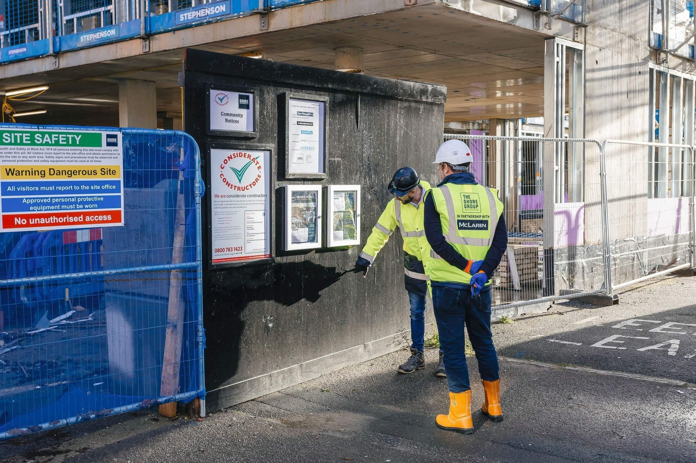 Two construction workers in safety gear examine a bulletin board on a building site, surrounded by safety signs and fencing.