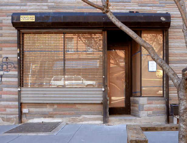 A storefront with a dark brown awning and a window with tan horizontal blinds. The building facade is made of horizontal brick or stone panels in beige and reddish-brown tones. A tree branch extends over the awning. A dark gray metal grate is visible on the sidewalk in front of the store. A small portion of a dark-colored object is visible to the right of the building on the right side of the image.