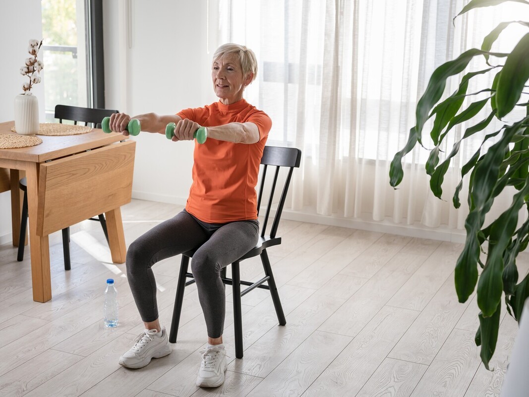 senior woman doing chair exercises to burn calories in her kitchen using dumbbells