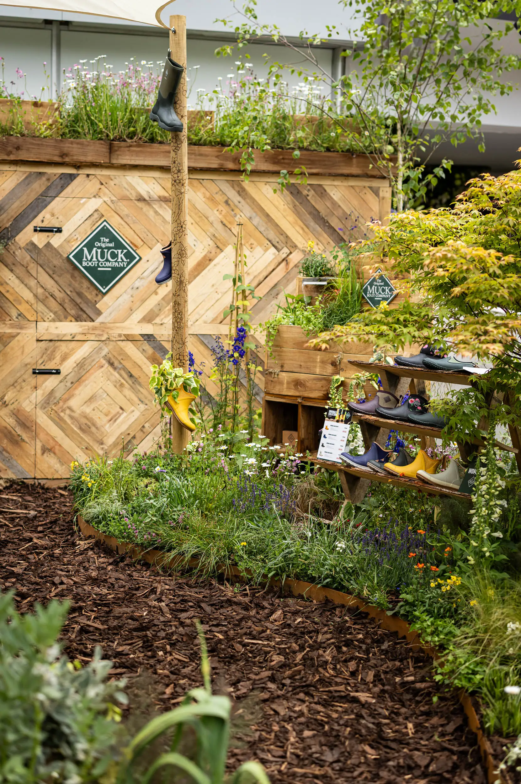 A garden scene featuring lush greenery, mulch, and a small structure in the background. Natural light illuminates the area.