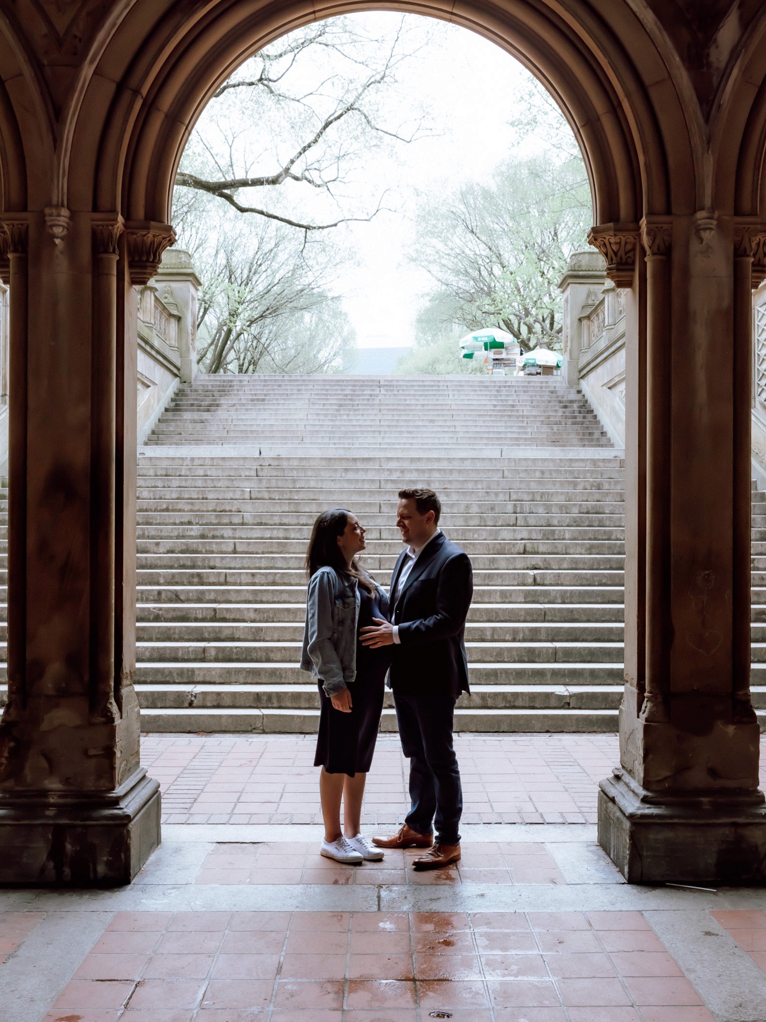 Glowing expecting couple smiling at the camera while cradling her baby bump under the ornate archway at Bethesda Terrace in Central Park, NYC — joyful, natural maternity photography by Lizz Spano Photography, New York City maternity photographer.
