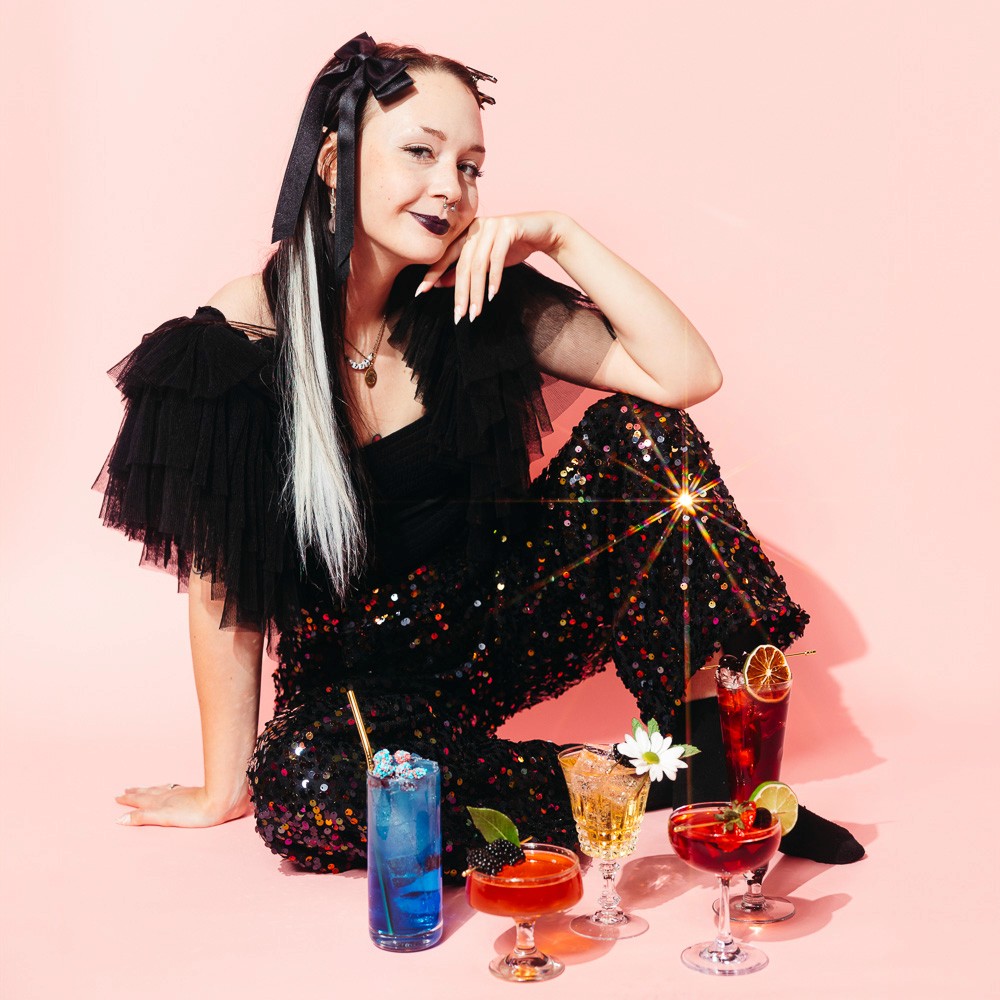 Sophie Liffick, owner of So Curious Co, poses in a sparkly suit with her non-alcoholic drinks on a pink background