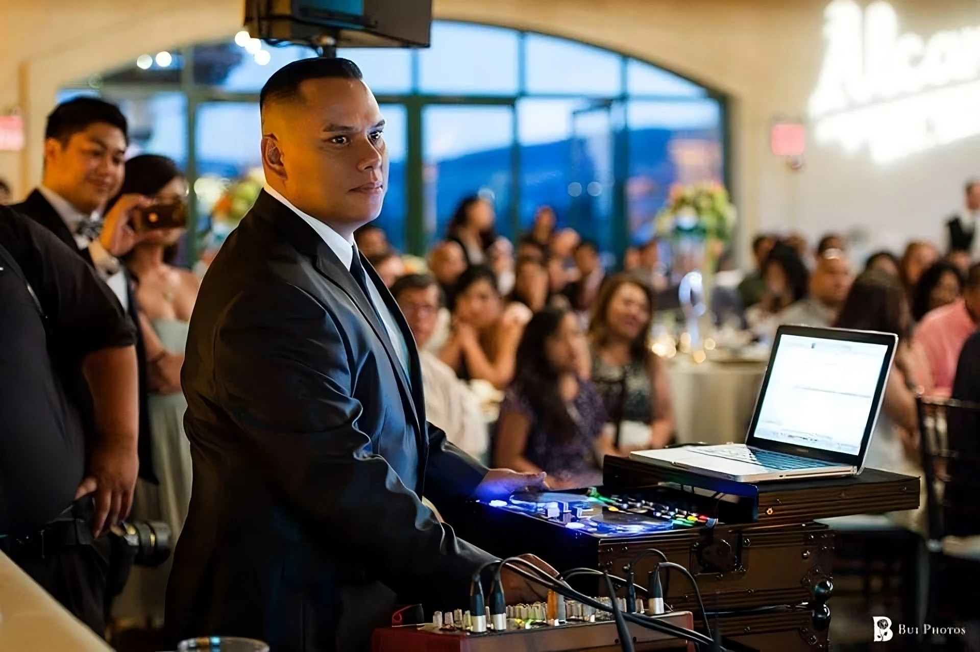 DJ Aaron in a black suit, smiling behind his DJ equipment at an event.
