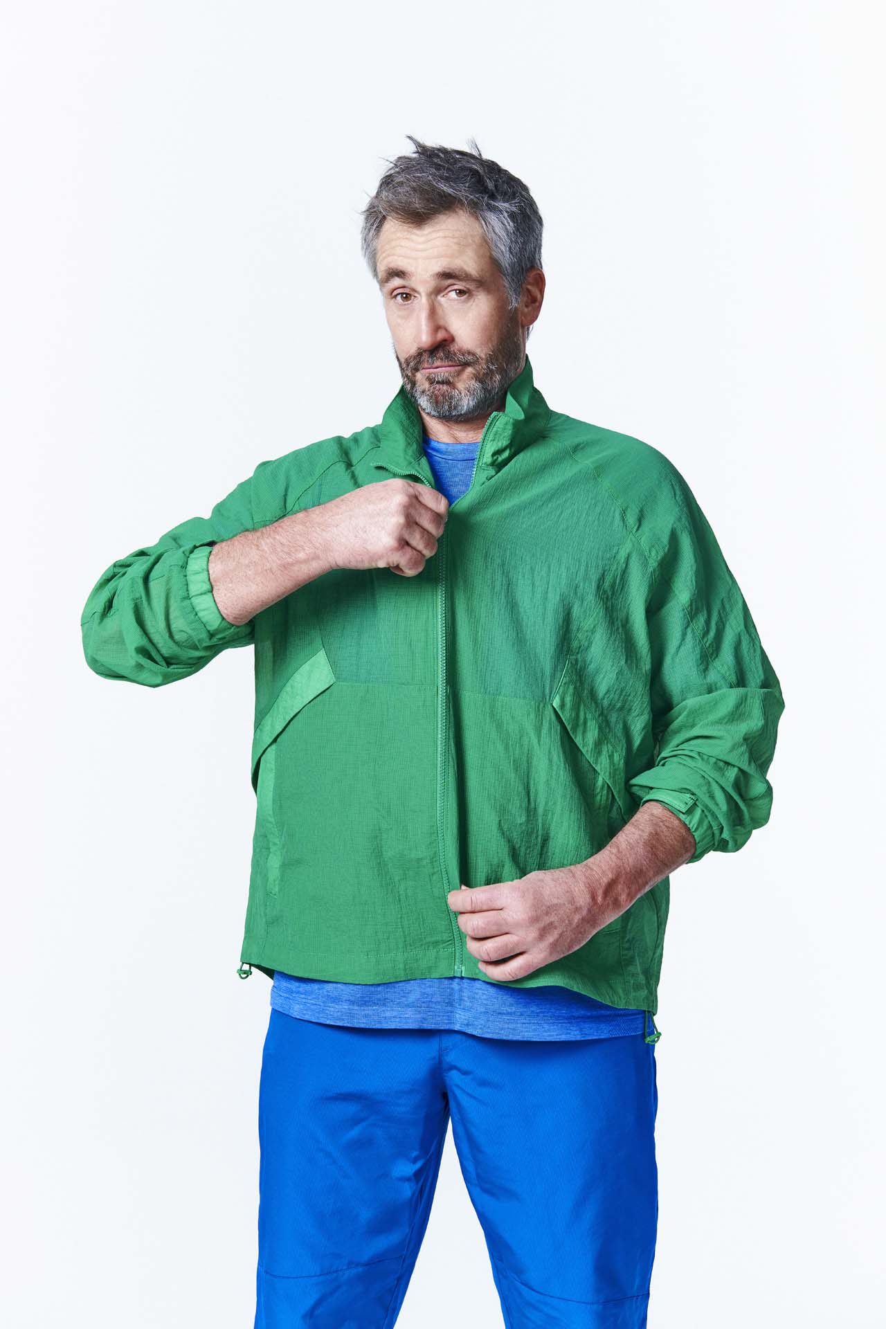 A person wearing a green jacket and blue pants adjusts their collar, with a plain light background.