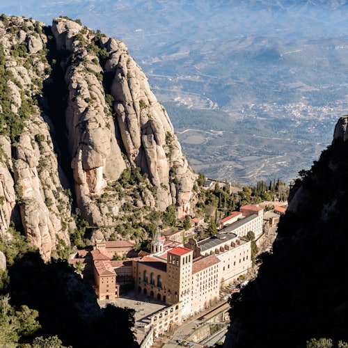Large monastery nestled between tall, rugged mountains with a valley and distant landscape in the background.