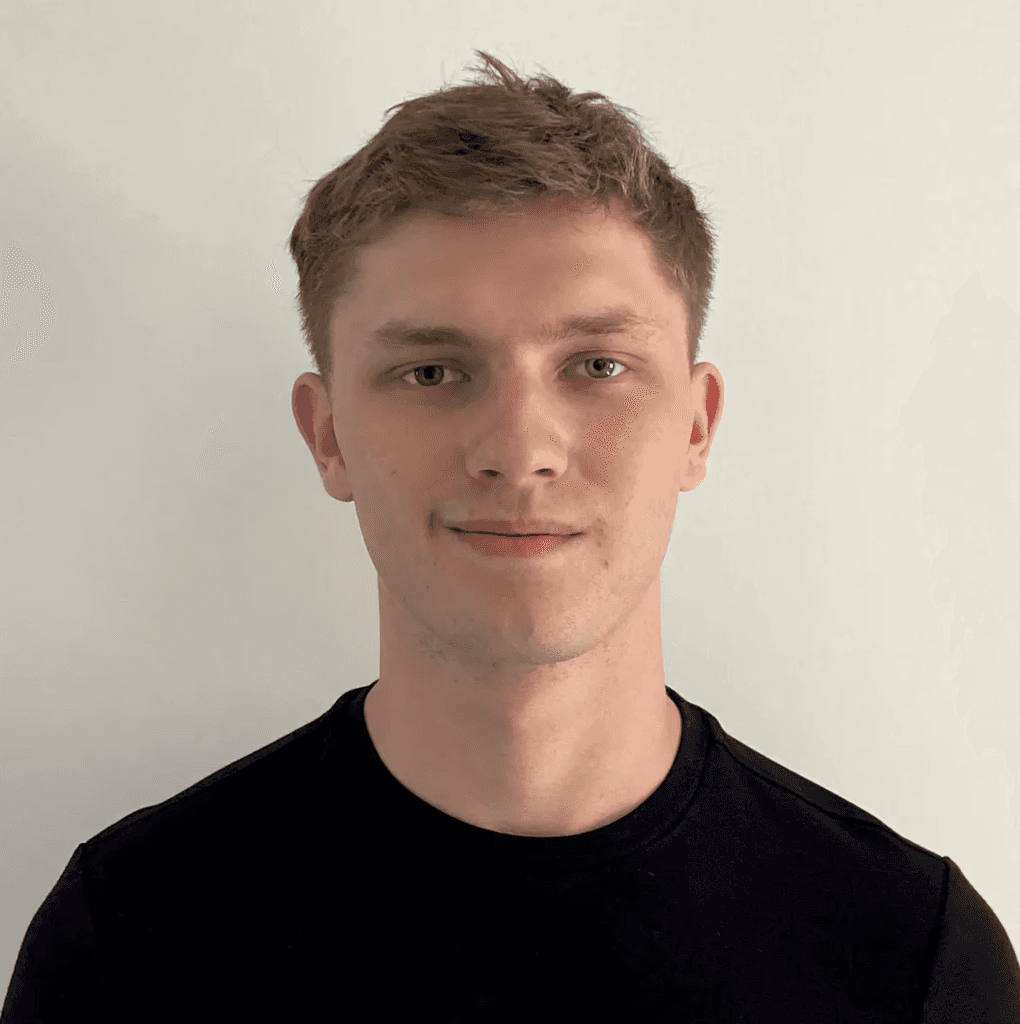 A smiling man with short brown hair in a dark blazer and white shirt against a light gray background. His expression conveys warmth and friendliness.