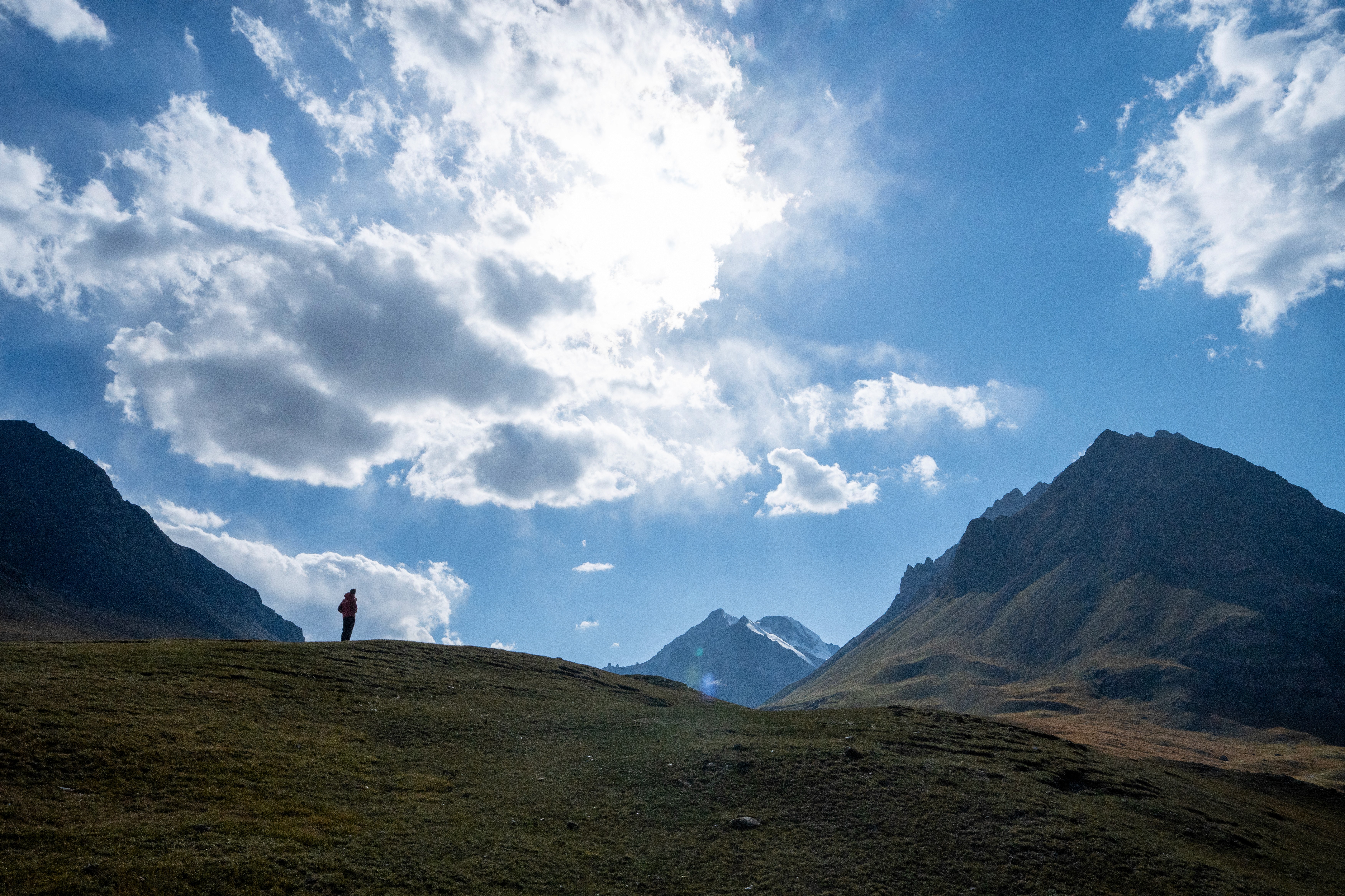 A lone figure on a grassy ridge beneath mountain peaks and a bright clouded sky