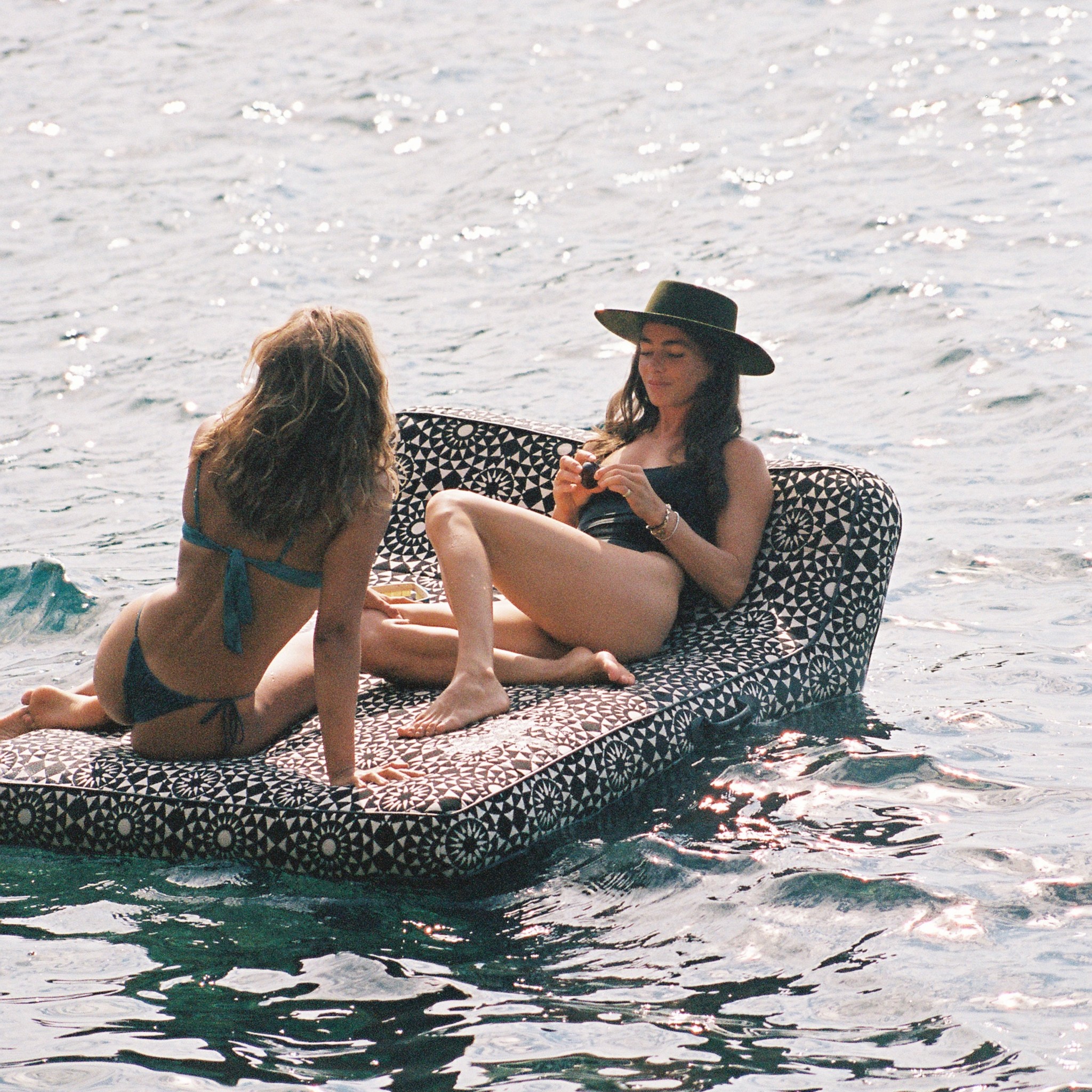 Two women lounging on a luxury black-and-white geometric pool float in the ocean eating figs.