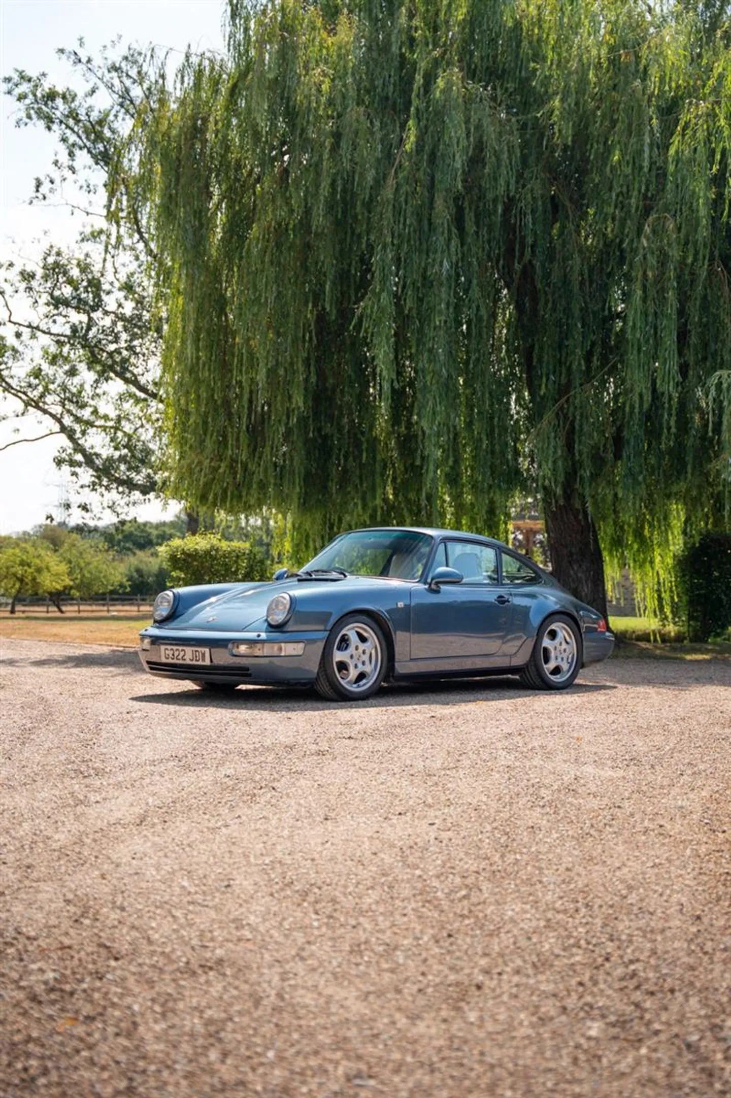 1990 Porsche 964 Carrera 2 in Baltic Blue Metallic, front three-quarter view beneath a willow tree