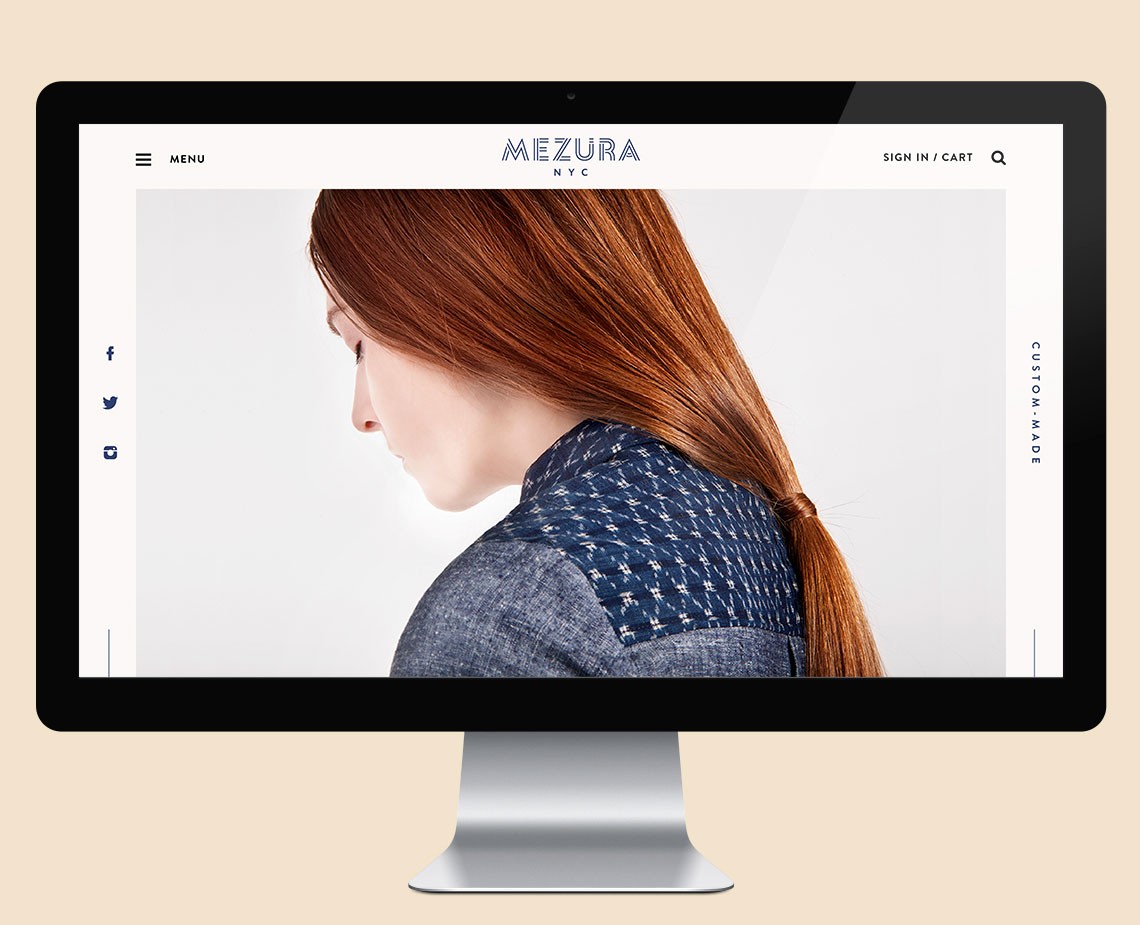 A computer monitor displays a website for Mezura NYC. The screen shows a woman with long reddish-brown hair, viewed from the side, wearing a dark blue sweater with a textured pattern. The background is light gray, and the website includes navigation links for "MENU," "SIGN IN / CART," and "CUSTOM MADE."