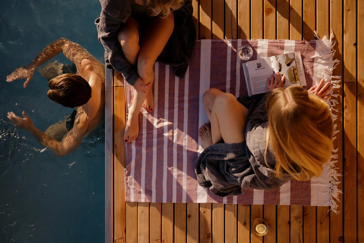 Zwei Frauen lesen auf einem gestreiften Tuch am Pool, während ein Mann seine Bahnen zieht