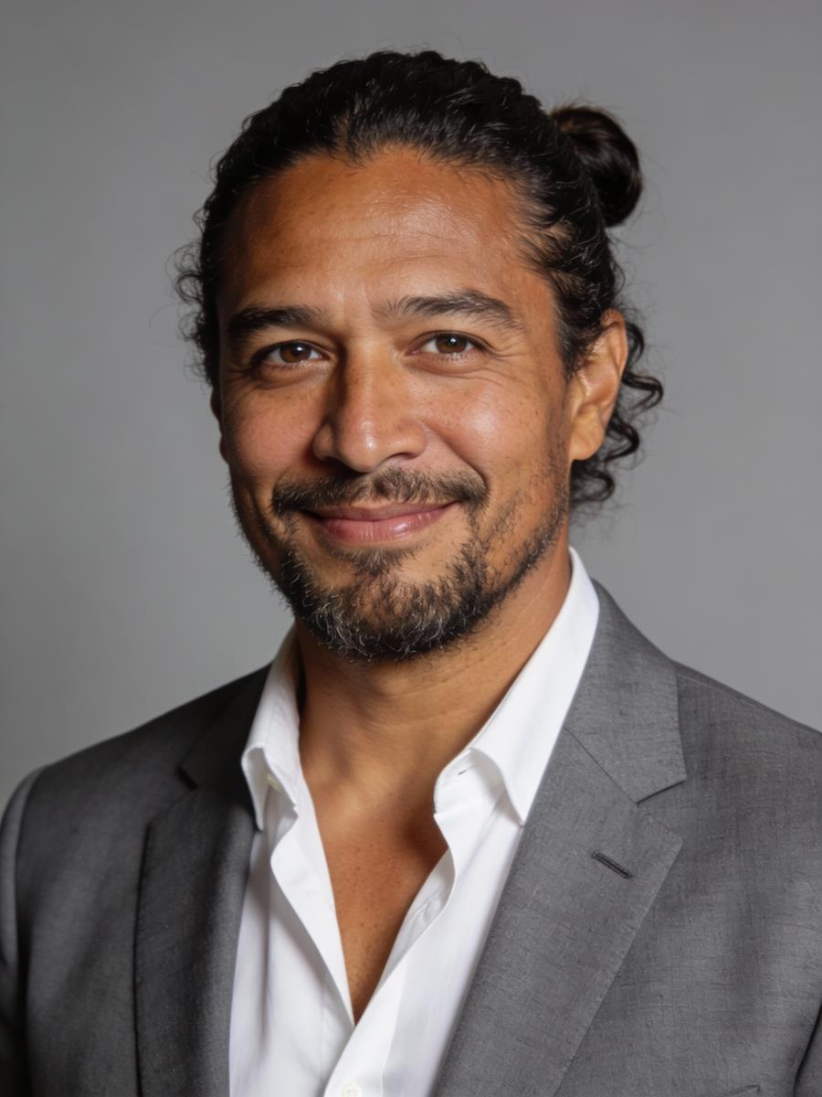 Smiling man with a beard, wearing a grey suit and white shirt, has dark hair tied back. Gray background gives a professional and approachable vibe.