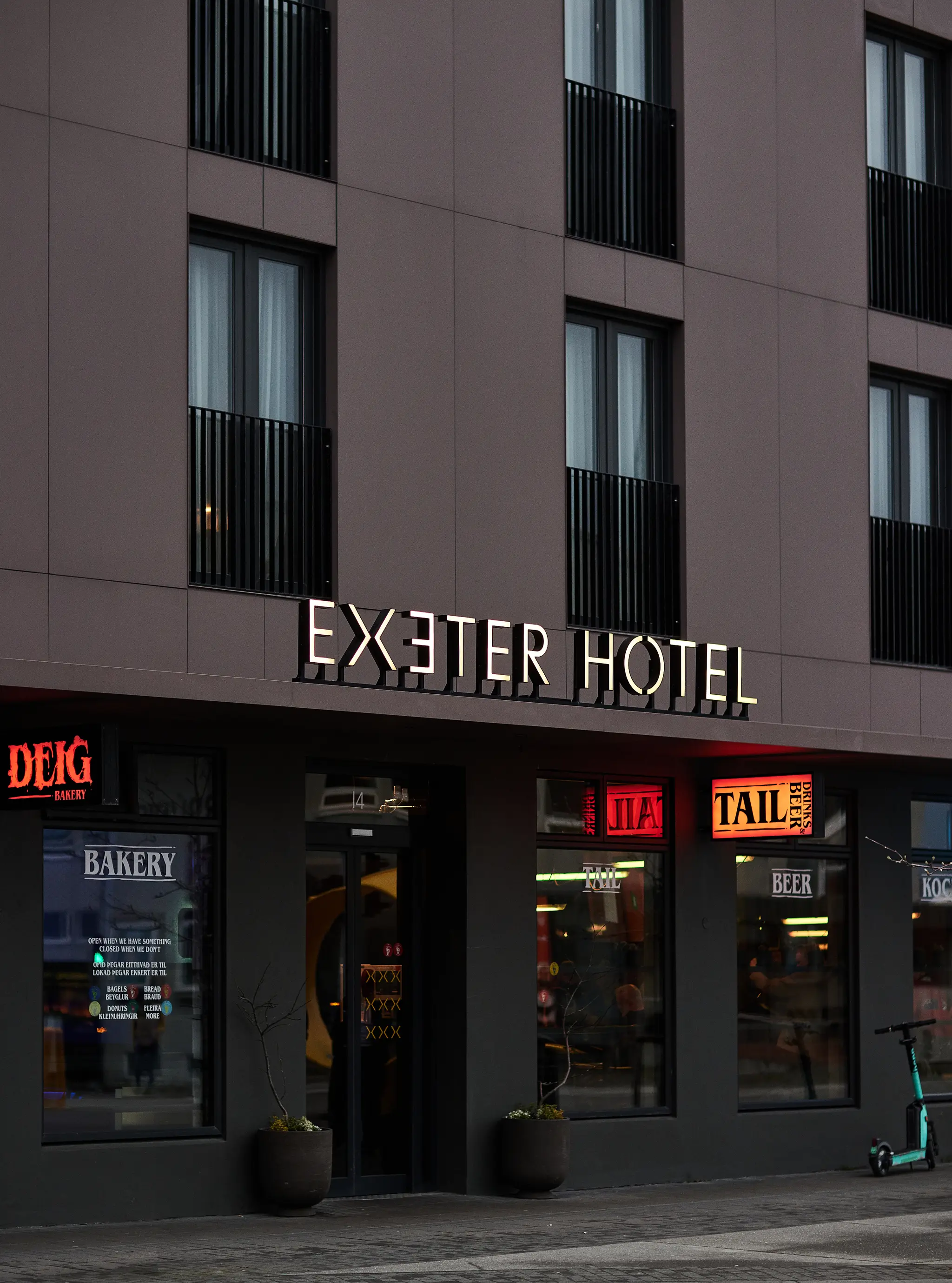 Exeter Hotel’s dark façade at dusk, with illuminated hotel lettering above the Deig bakery and Tail bar signs