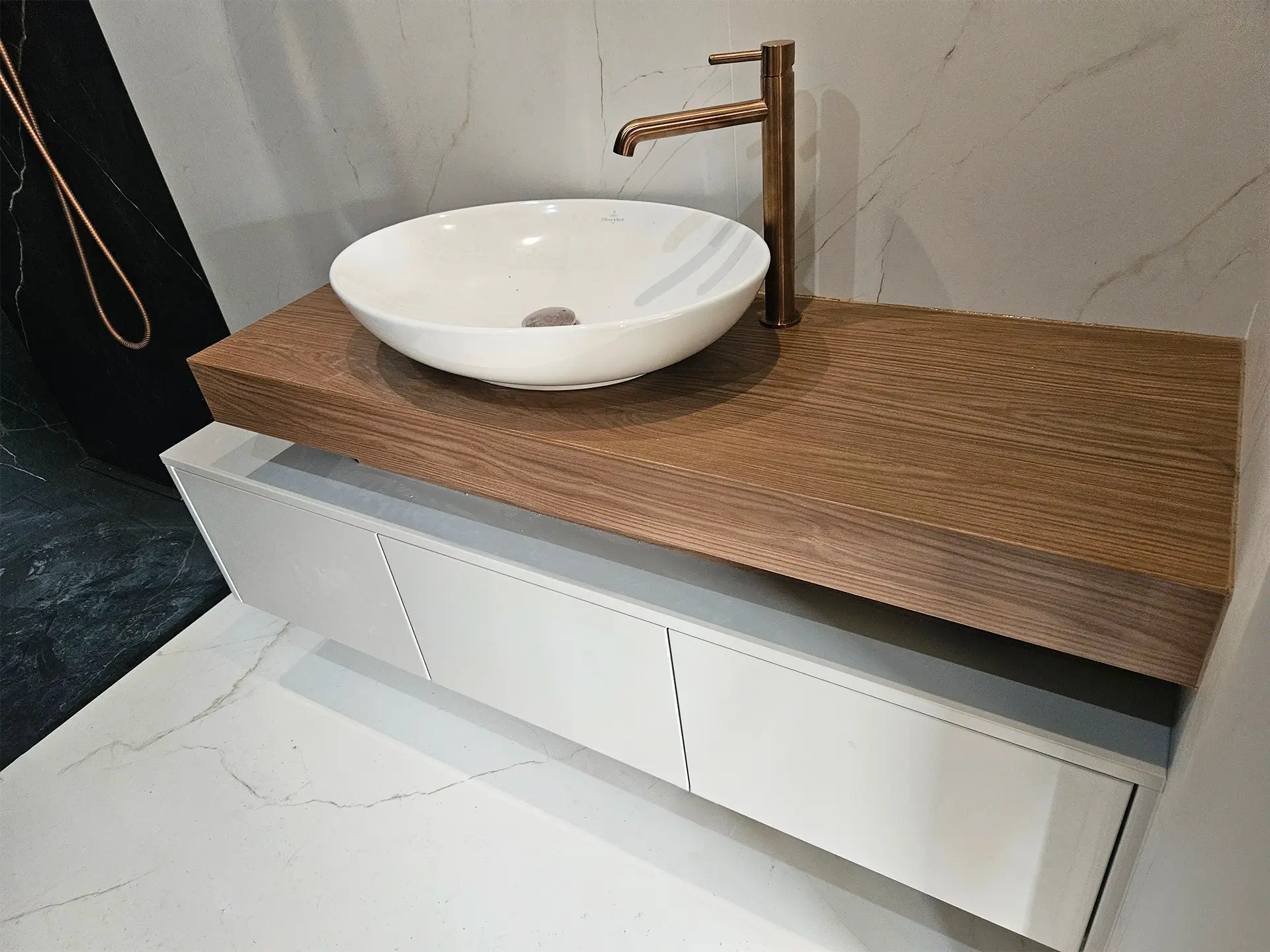 Podwieszana szafka łazienkowa z białymi frontami, grubym drewnianym blatem i miedzianą baterią.