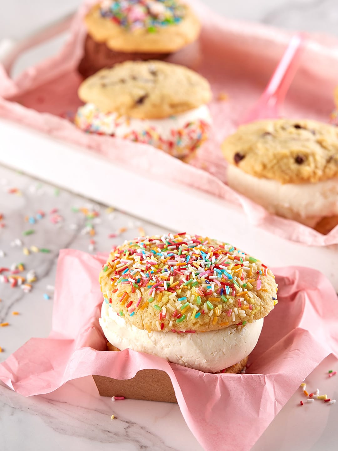 Vanilla ice cream sandwich with sprinkle cookies on pink napkin with more sandwiches in the blurred background.