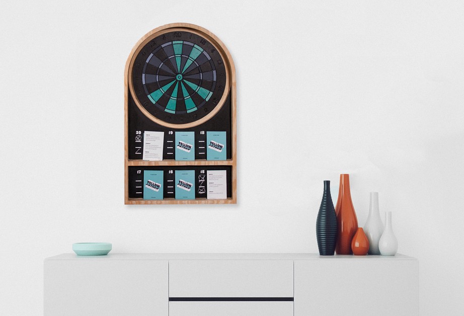 A circular wooden dartboard with a teal center and black outer ring is mounted on a white wall. Below it, a wooden frame displays numbered slots with small cards inside. The frame is mounted above a white cabinet with a light teal bowl on top and four vases of varying colors and sizes placed to the right.
