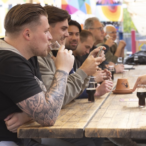 Group of people sitting at a long wooden table, drinking and socializing. Man in focus has tattoos, some look at phones. Decorated background.