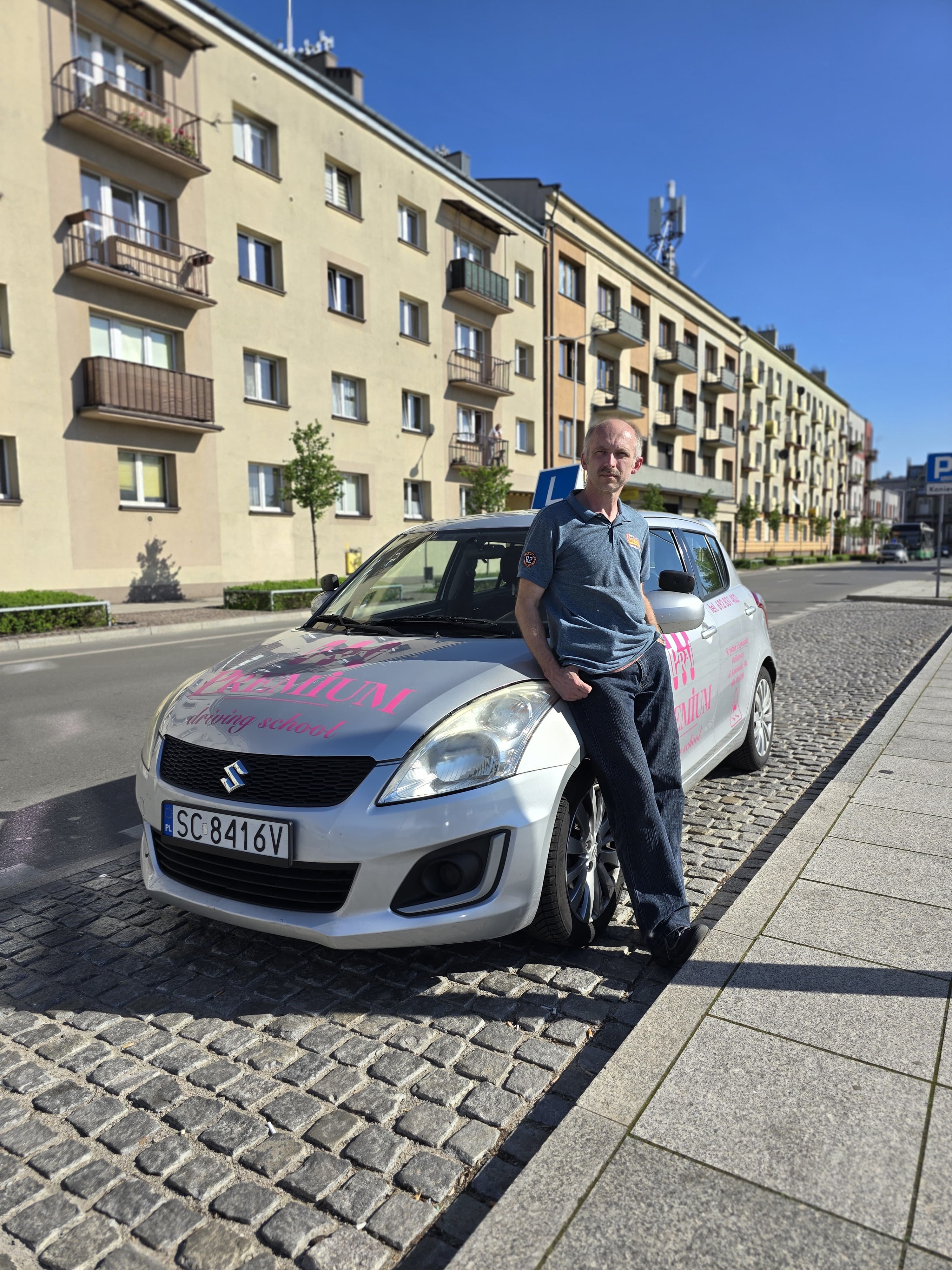 Piotrek Oberski – Instruktor przy srebrnym Suzuki Swift oznaczonym logo szkoły jazdy.
