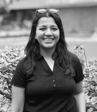 A smiling woman with her arms crossed, standing against a dark green background. She has long, dark hair.