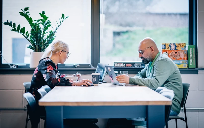 Two people sit across from each other at a long table in a bright, modern office space, working on laptops. One person is writing in a notebook beside a coffee cup, while the other focuses on their screen. Board games are stacked on a window ledge in the background, and a large plant and window provide natural light, creating a collaborative and informal working environment.
