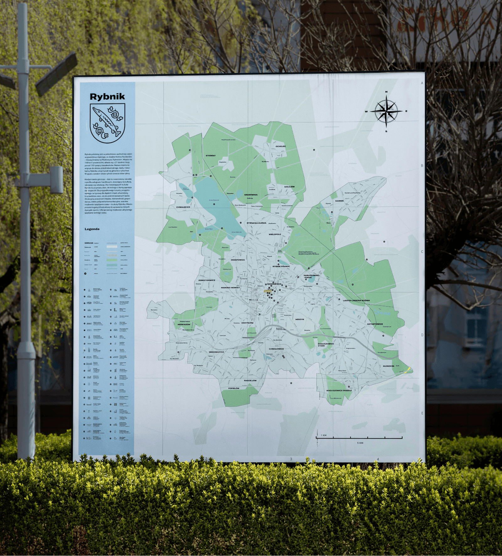 Duża tablica z mapą miasta, nagłówkiem „Rybnik”, herbem i legendą, stojąca za żywopłotem obok latarni.