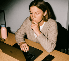A woman sits at a desk with a laptop, deep in thought, holding her chin, with a bottle beside her.