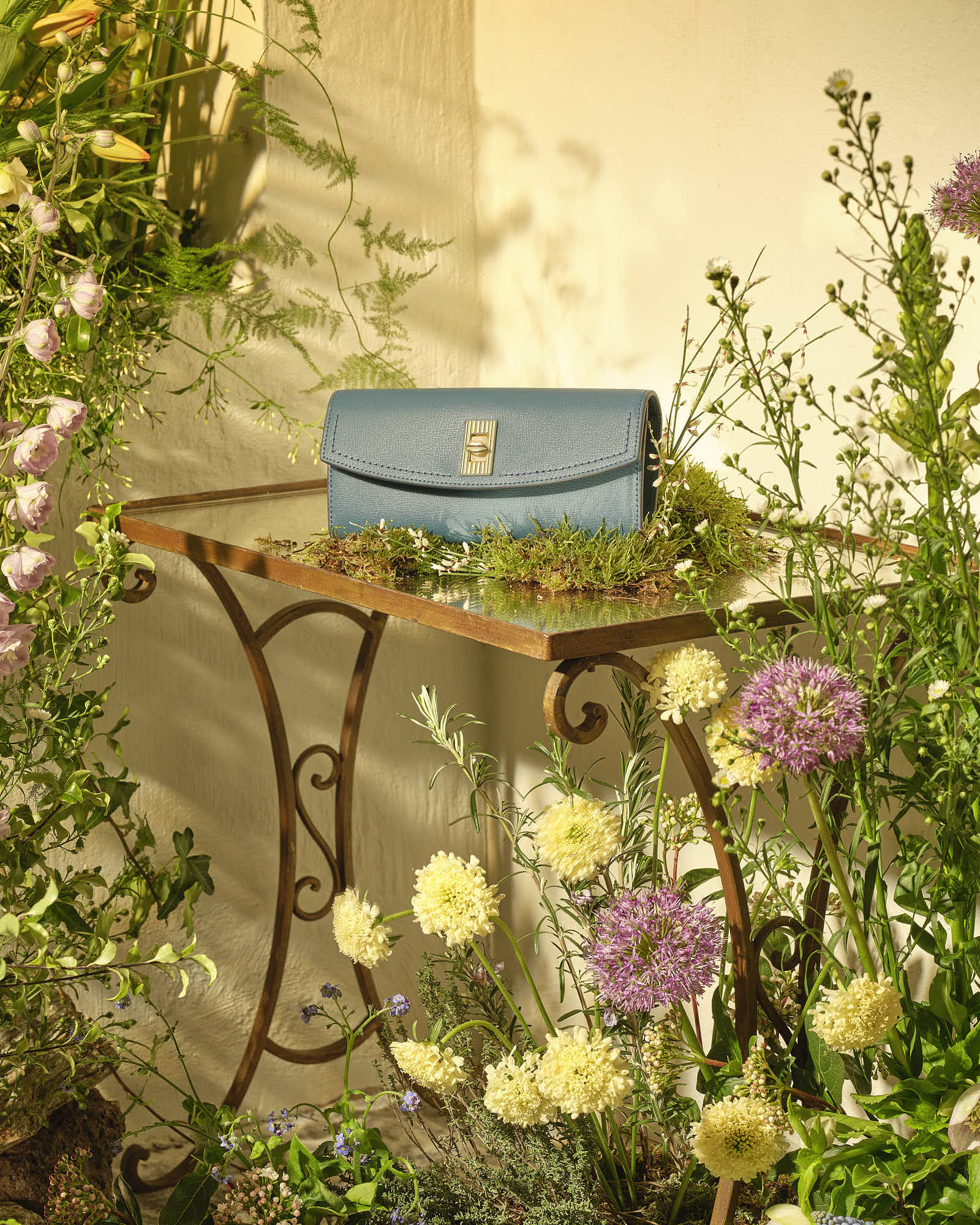 Blue clutch purse on a rustic table surrounded by vibrant flowers in sunlight.