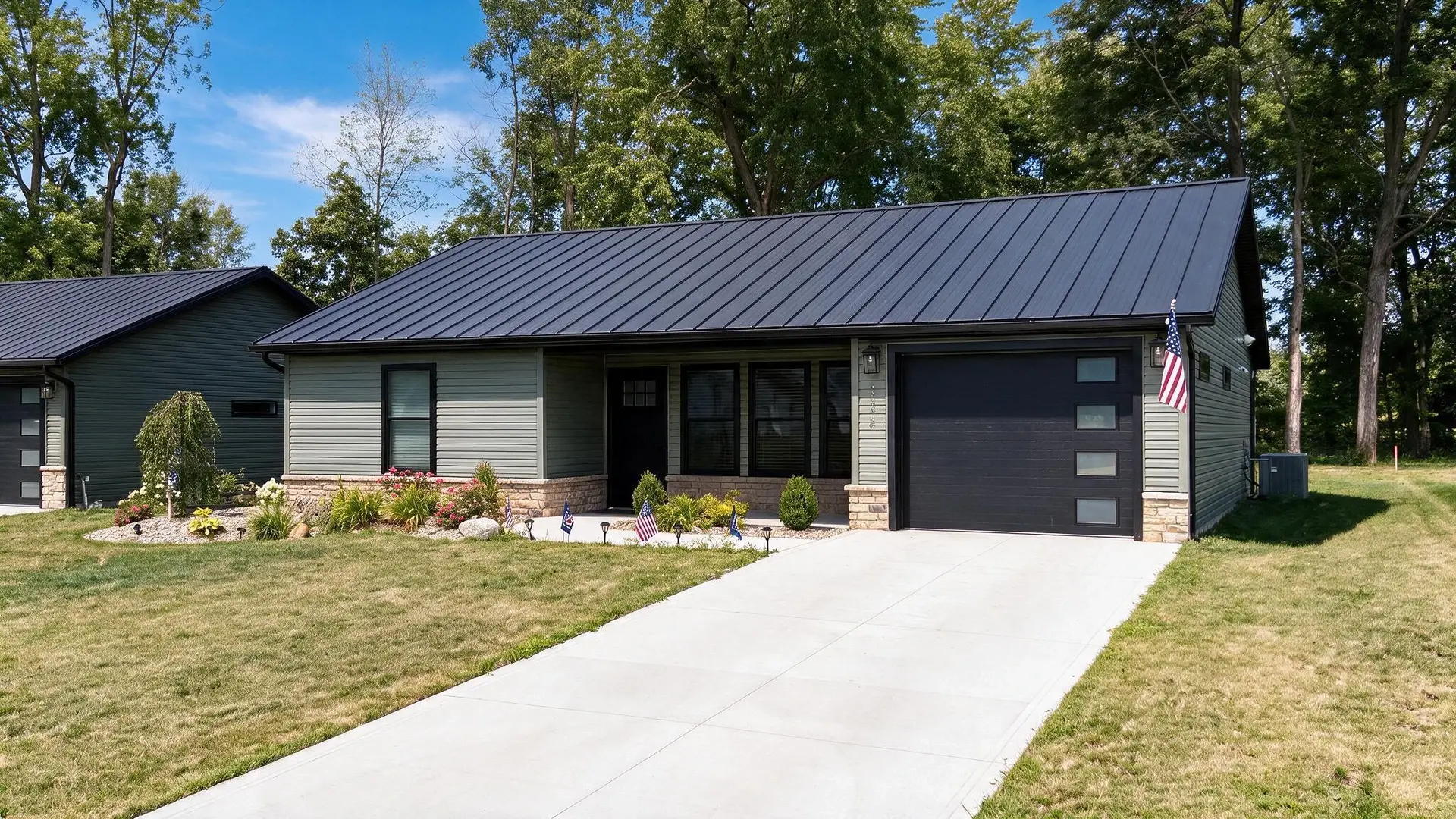A finished green-sided ranch home with a black metal roof, planted front beds, an American flag by the door and a long concrete driveway