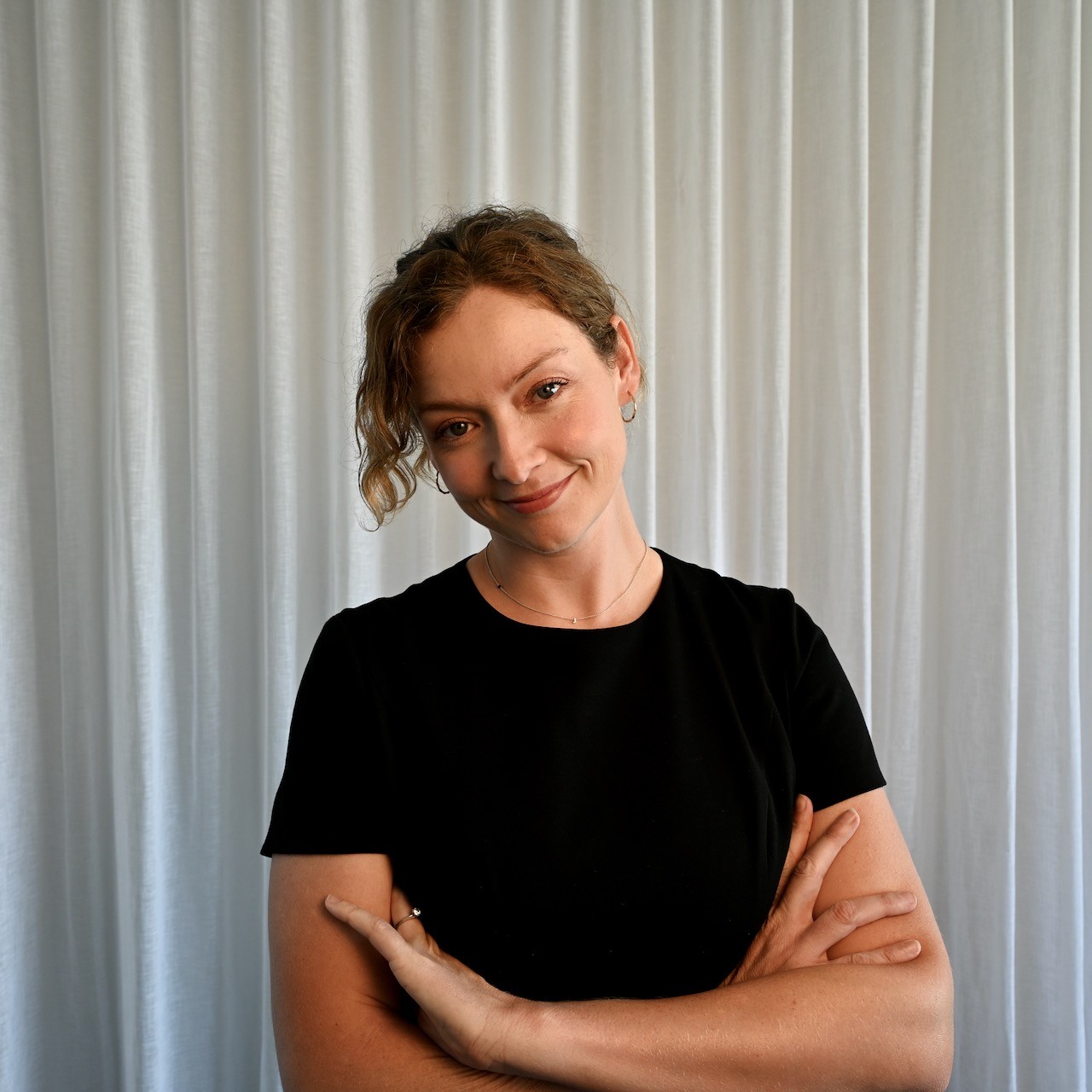 A smiling man with curly hair is sitting at a desk, looking at the camera, with a laptop and a lamp in the background.