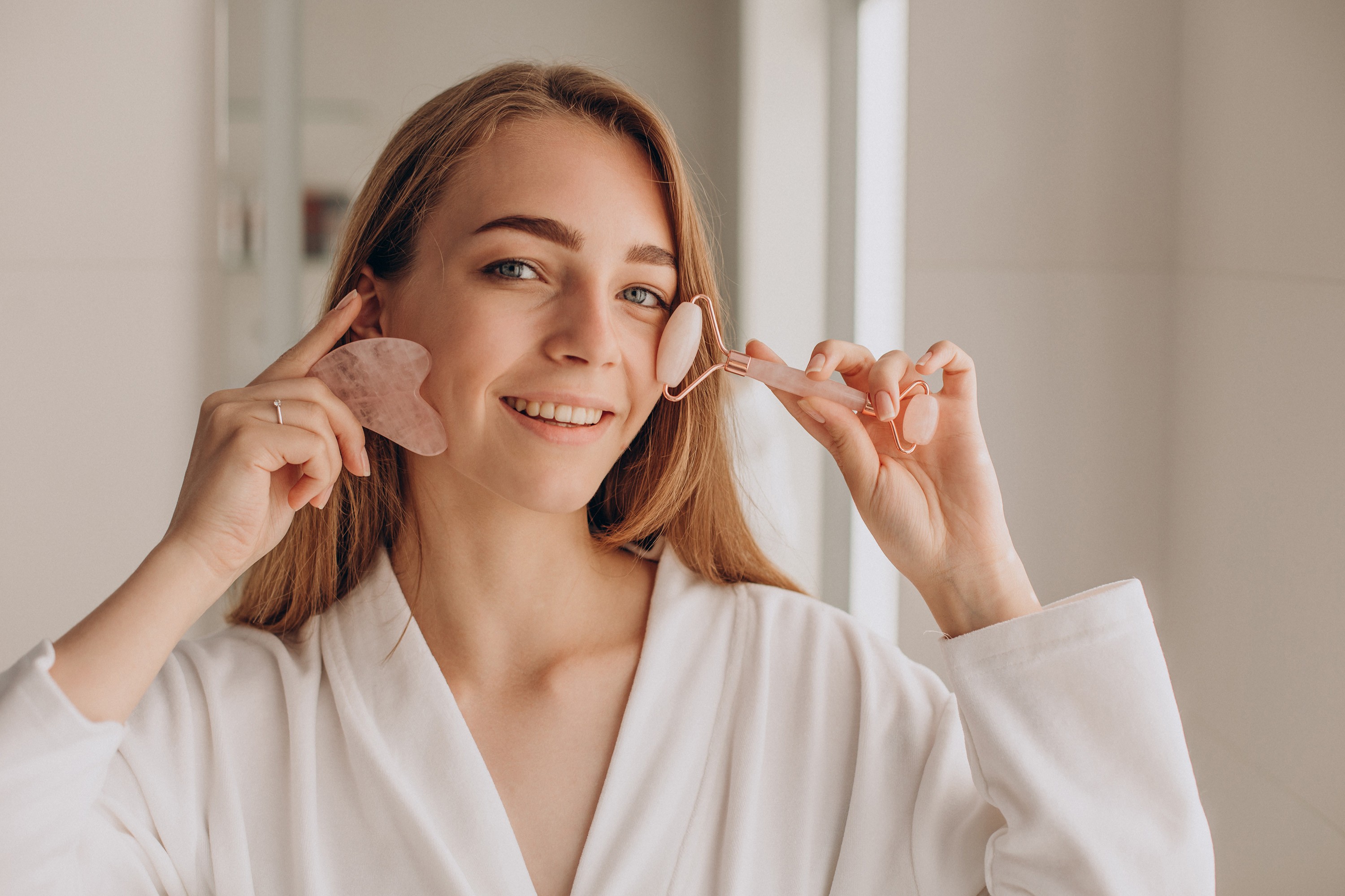 Smiling woman using a rose quartz Gua Sha stone and face roller for a facial massage