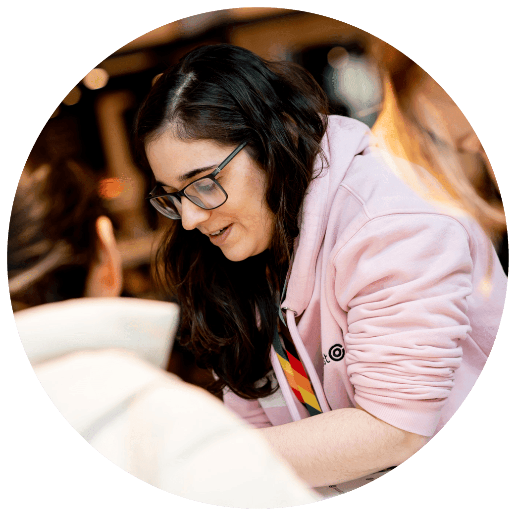 Person wearing a pink hoodie with a rainbow lanyard, leaning forward while engaging with others at a table.