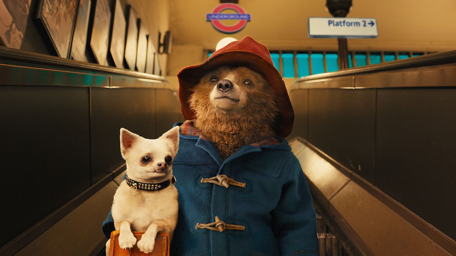 Paddington Bear in his blue coat and red hat at a London Underground station with a small dog, in 'Paddington 3'.