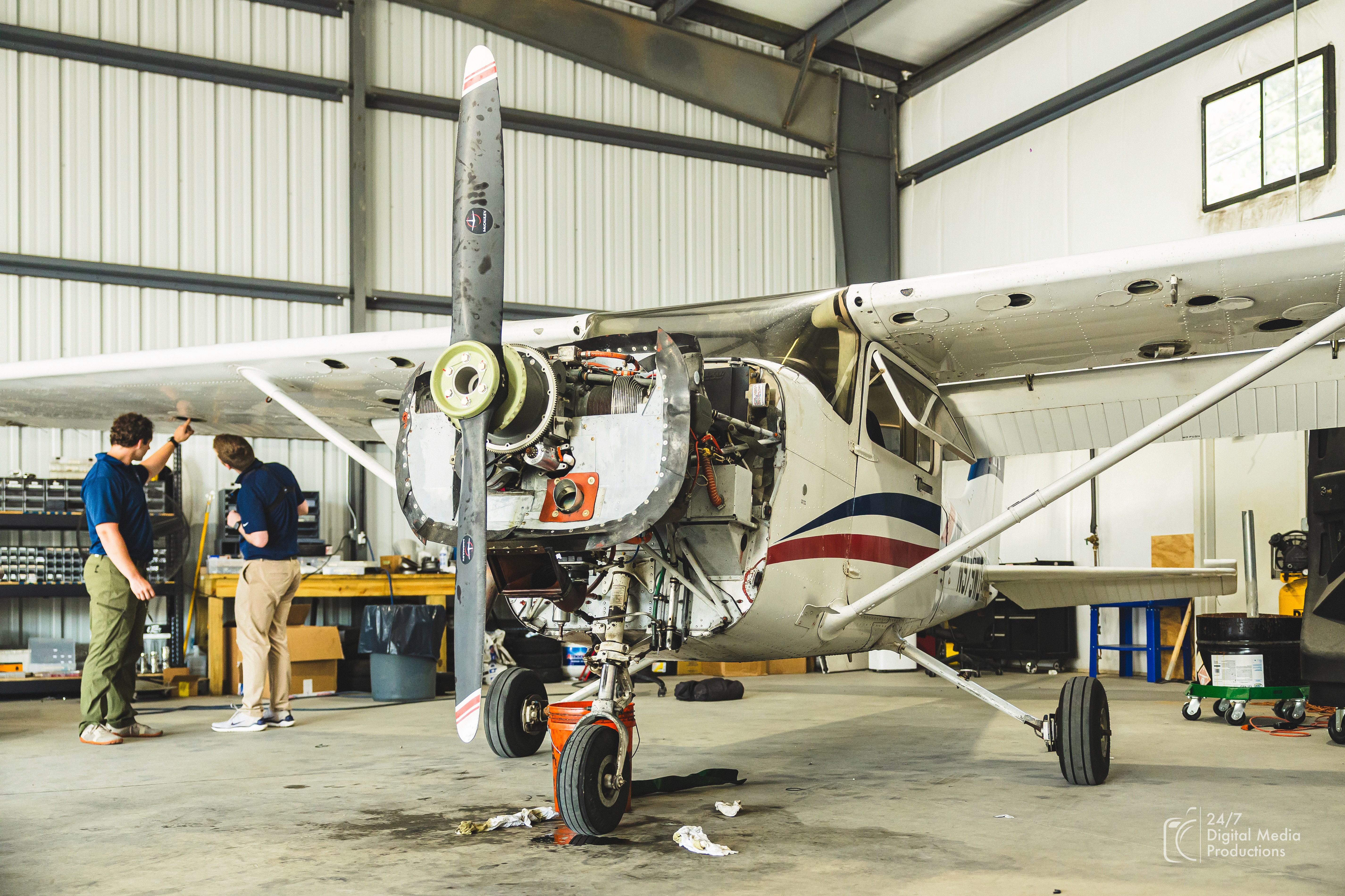 A mechanic in blue overalls is working on the engine of a small airplane, which is exposed with many pipes, mechanical parts, and the propeller visible.