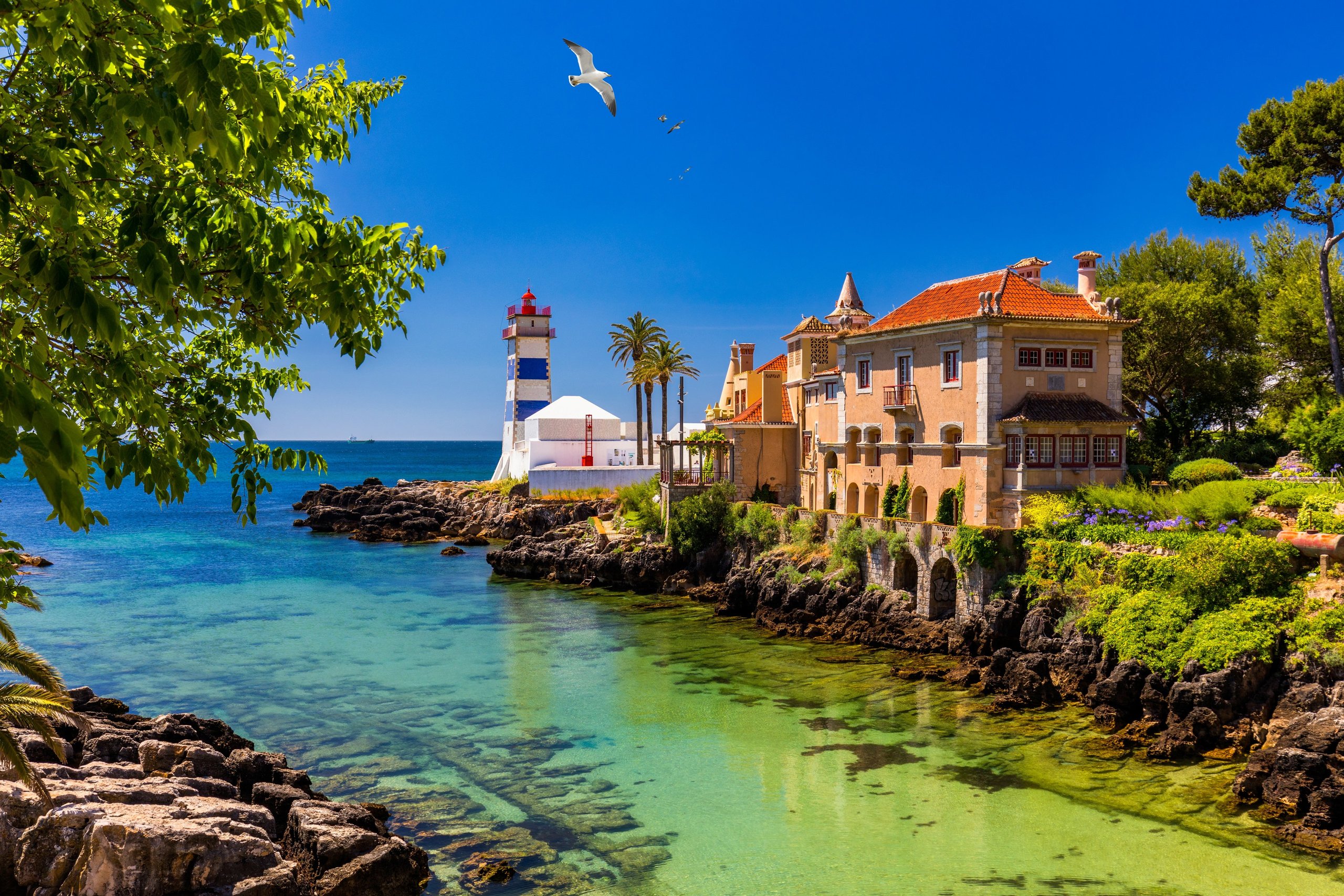 The blue-and-white striped Santa Marta Lighthouse in Cascais and the adjacent Casa de Santa Maria (a Portuguese Romantic-era manor with an orange-red tile roof), green-tinged shallow water in the foreground, a seagull overhead.
