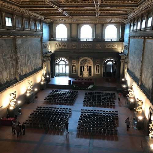 Grande sala con soffitto decorato, ampi murales e file di sedie disposte per un evento. Alcune persone sono in piedi e conversano.