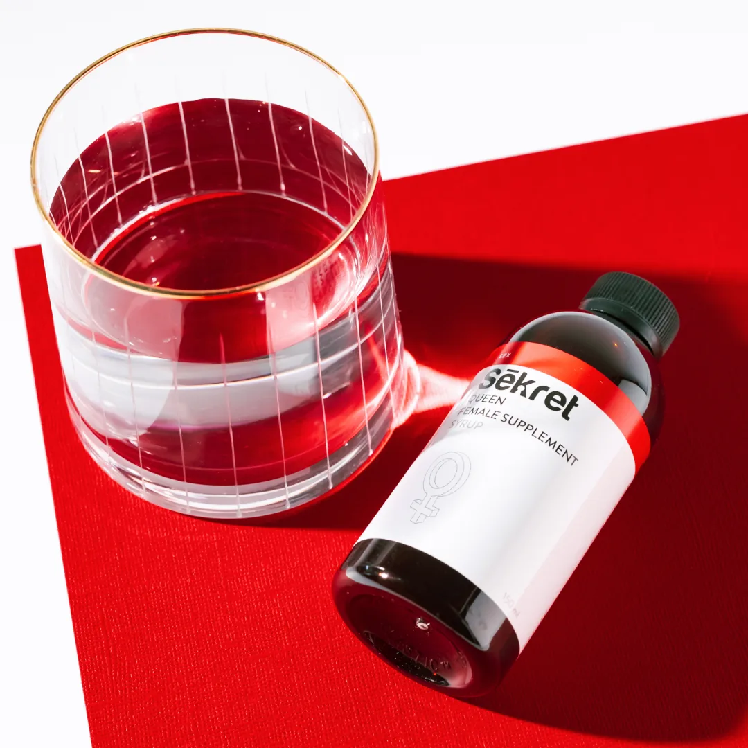 A bottle of Sēkret Queen syrup lying beside a glass of red drink on a red card