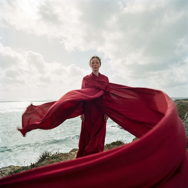 A poised person in flowing crimson robes stands on a rugged seaside cliff, wind sweeping the fabric over the ocean beneath dramatic clouds.