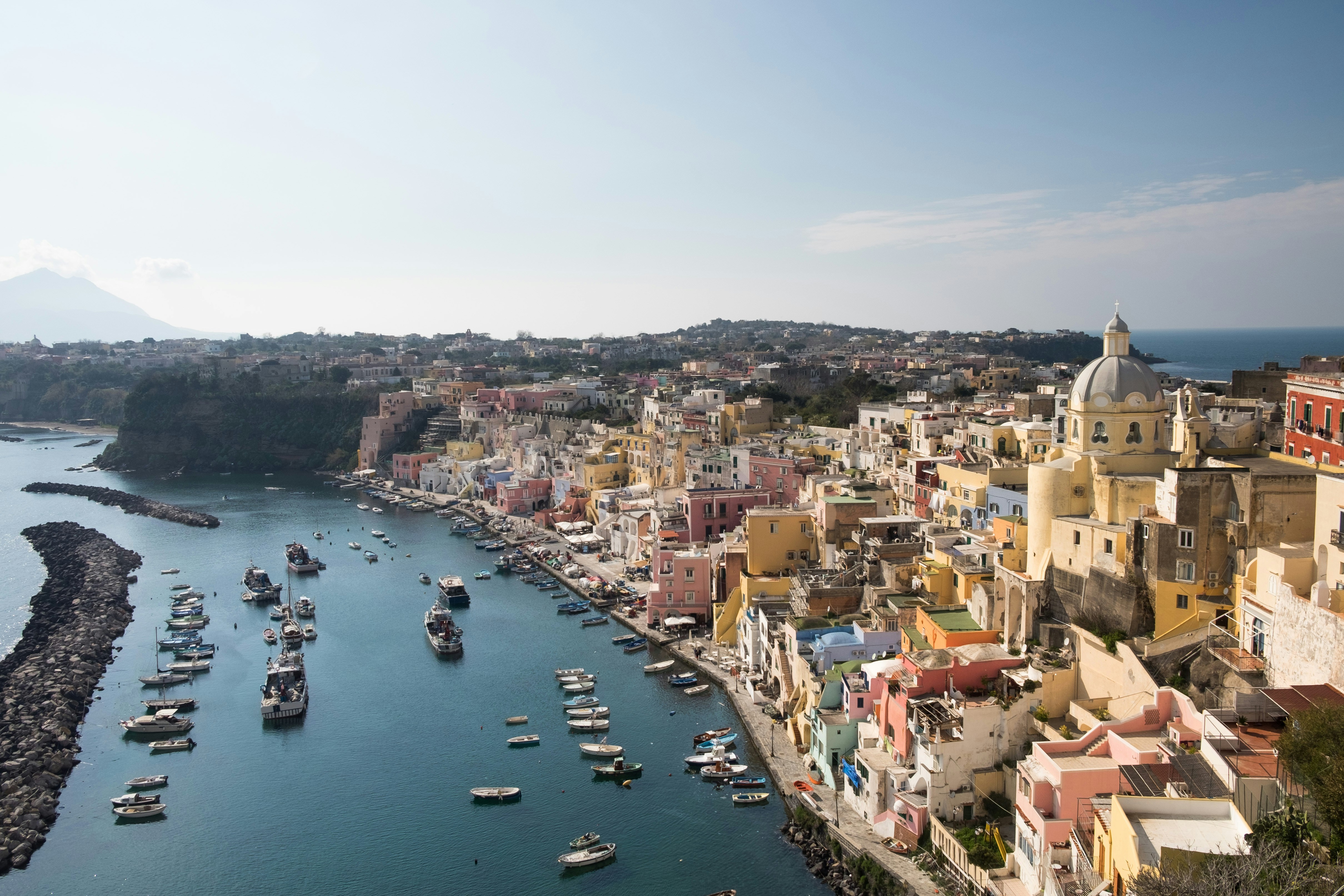 Vista panoramica di Procida, un'isola pittoresca della Campania, con case colorate lungo la costa, barche ormeggiate e il mare azzurro sotto un cielo limpido.