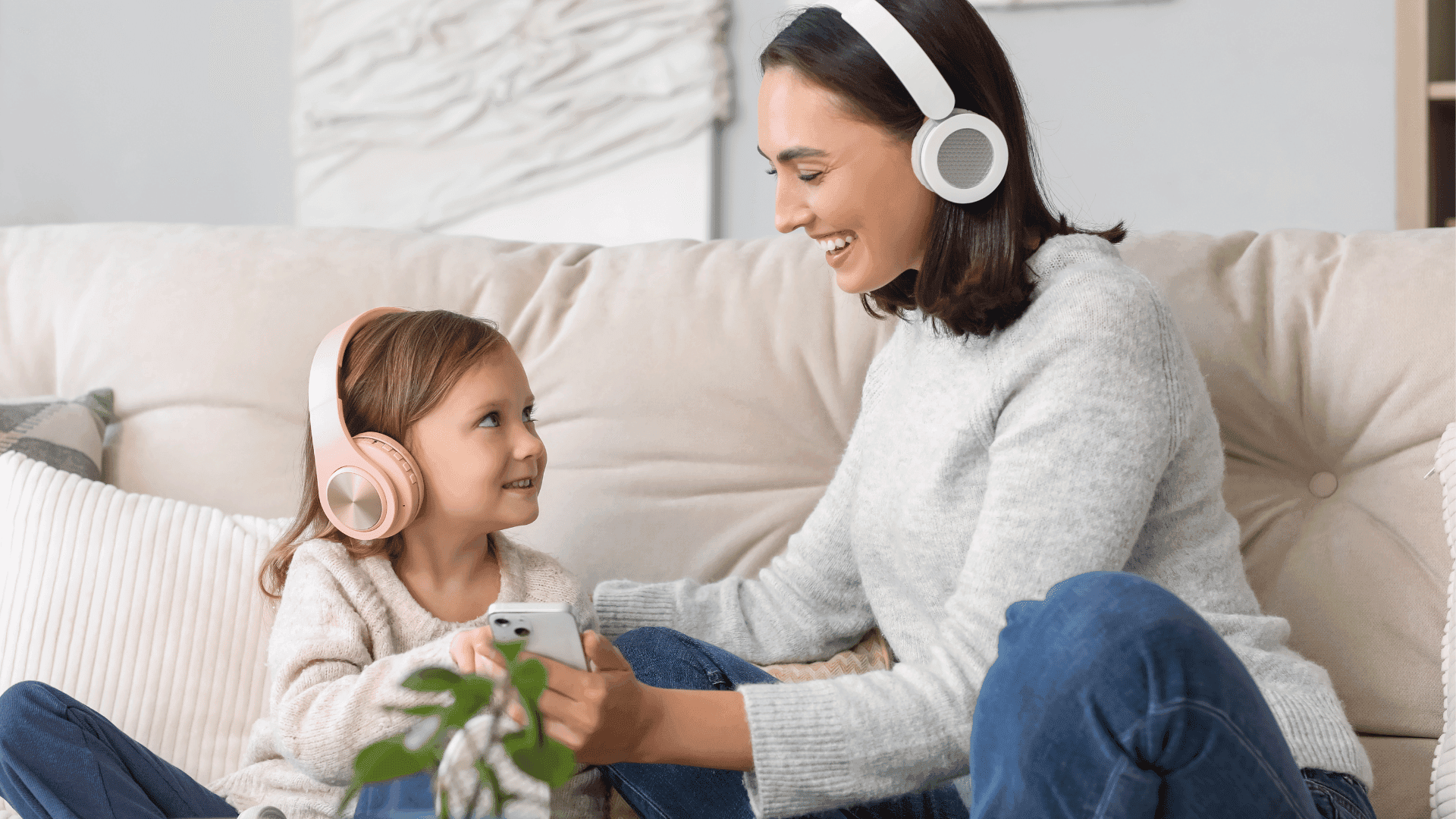 A mum and daughter sitting on a sofa, both wearing headphones, with the mum holding a phone as they listen to HushAway®’s Sound Sanctuary.