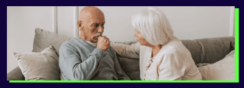 Elderly couple sitting together on sofa, one appearing unwell.