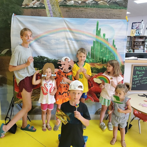 เด็กหกคนถ่ายรูปพร้อมพร็อพท่ามกลางฉากหลังธีมออซ ซึ่งประกอบด้วยสายรุ้งและเมืองมรกต