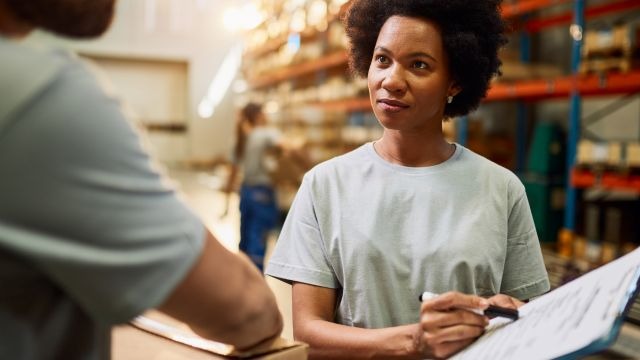 Em um depósito, mulher preta segurando prancheta e caneta enquanto observa homem à sua frente.